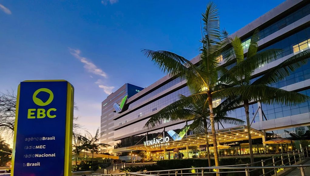 A imagem mostra, ao fundo, a fachada do edifício Venâncio Shopping, onde fica localizada a sede da Empresa Brasil de Comunicação, a EBC. Do lado esquerdo, em primeiro plano, uma placa vertical instalada na calçada com o logotipo da EBC sobre um fundo azul escuro. O logotipo é verde claro sobre um fundo azul escuro. Abaixo do logotipo, está escrito em letras brancas Agência Brasil, Rádio MEC, Radio Nacional e TV Brasil. Na legenda da foto, está o crédito do fotógrafo: Rafa Neddermeyer/Agência Brasil.