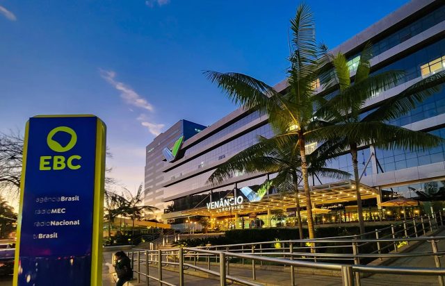 A imagem mostra, ao fundo, a fachada do edifício Venâncio Shopping, onde fica localizada a sede da Empresa Brasil de Comunicação, a EBC. Do lado esquerdo, em primeiro plano, uma placa vertical instalada na calçada com o logotipo da EBC sobre um fundo azul escuro. O logotipo é verde claro sobre um fundo azul escuro. Abaixo do logotipo, está escrito em letras brancas Agência Brasil, Rádio MEC, Radio Nacional e TV Brasil. Na legenda da foto, está o crédito do fotógrafo: Rafa Neddermeyer/Agência Brasil.